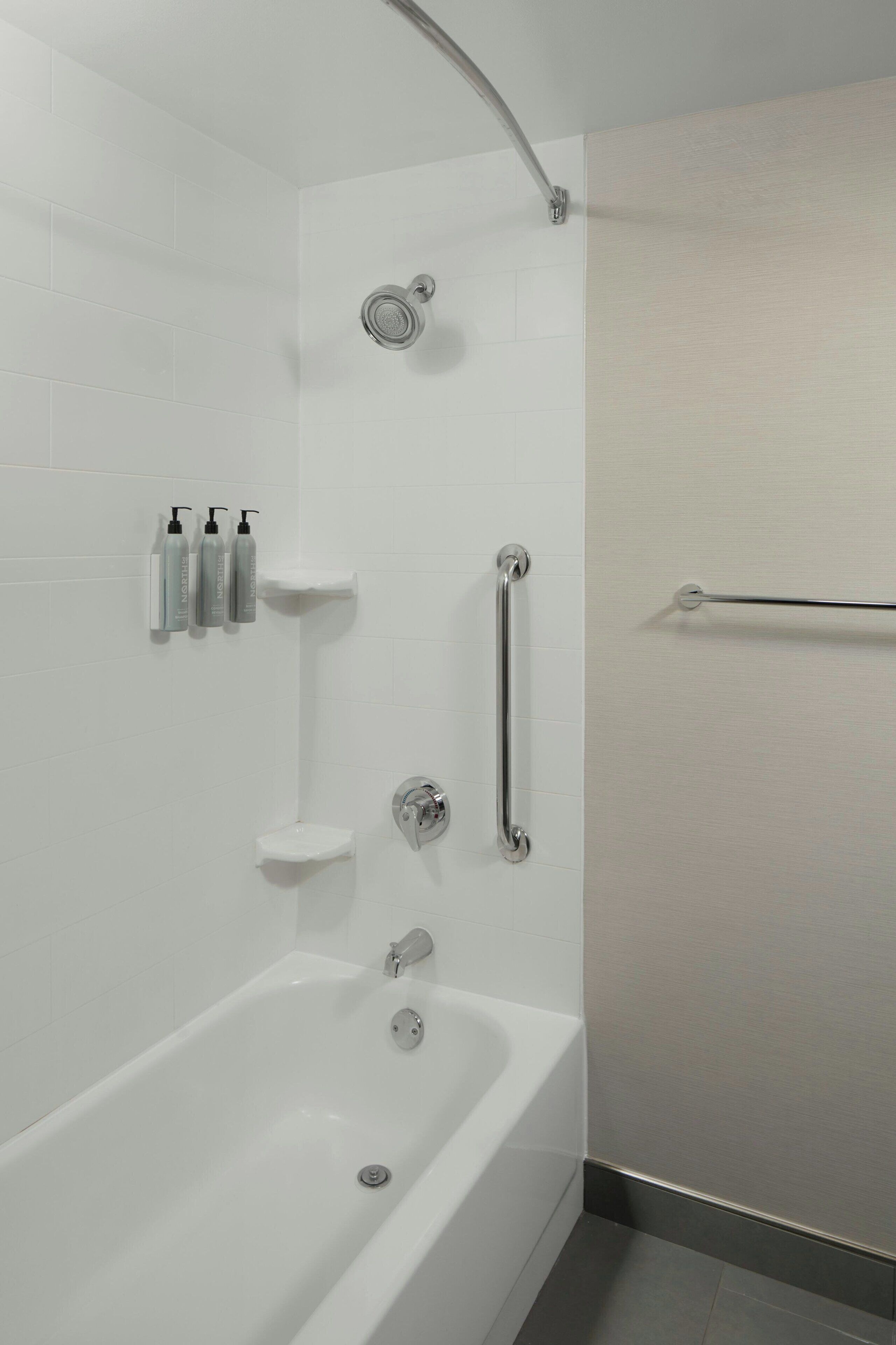 Une salle de bain moderne avec une baignoire, une pomme de douche, une barre d’appui et des articles de toilette rangés sur une étagère.