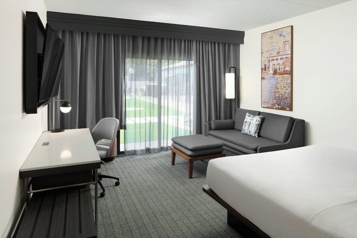 Habitación de hotel con cama, escritorio, silla, sofá y un amplio ventanal con cortinas.