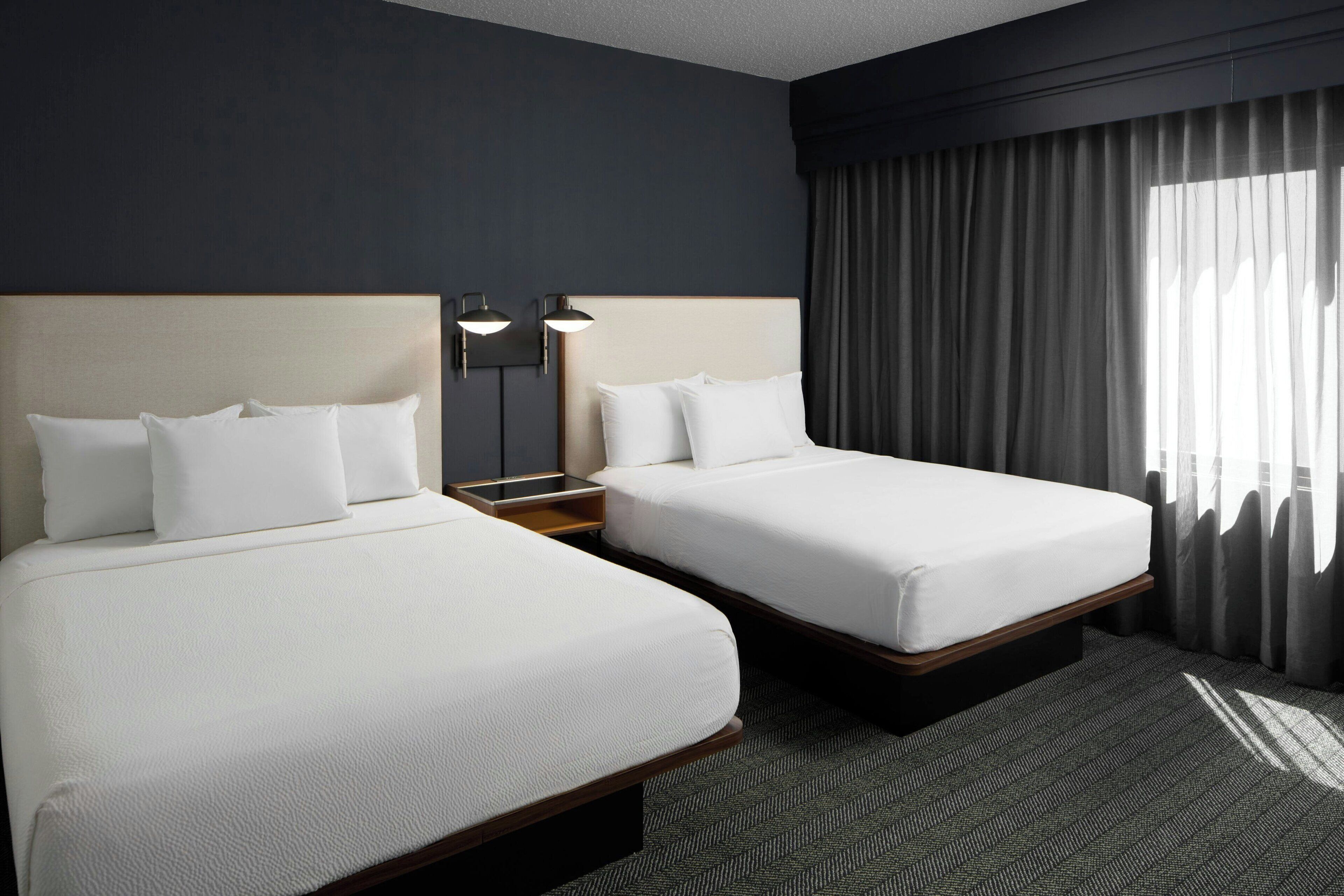 A hotel room with two beds, a dark wall, a window with curtains, and a headboard with a light fixture.