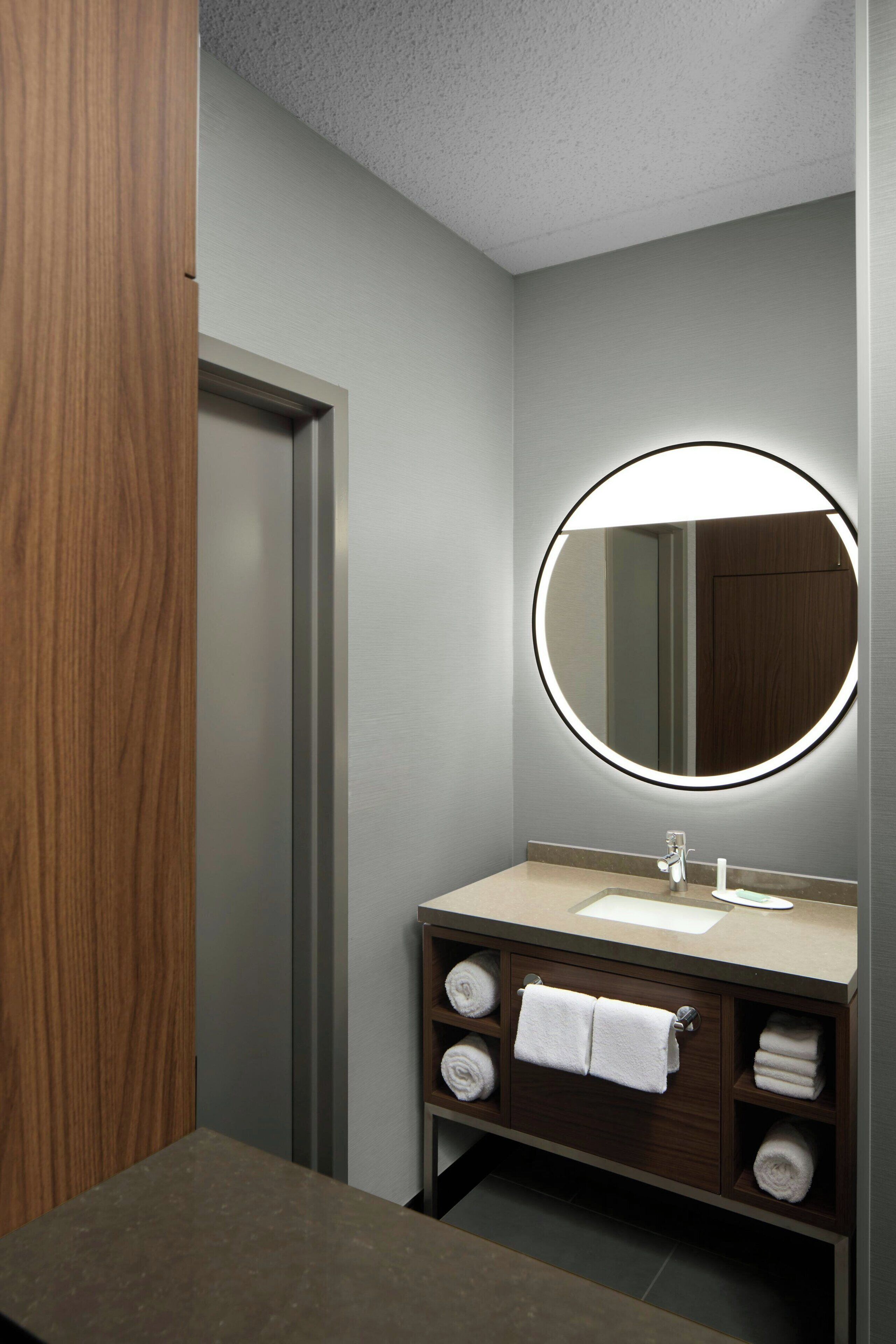 A modern bathroom with a wooden vanity, a round mirror with integrated lighting, and neatly folded towels.