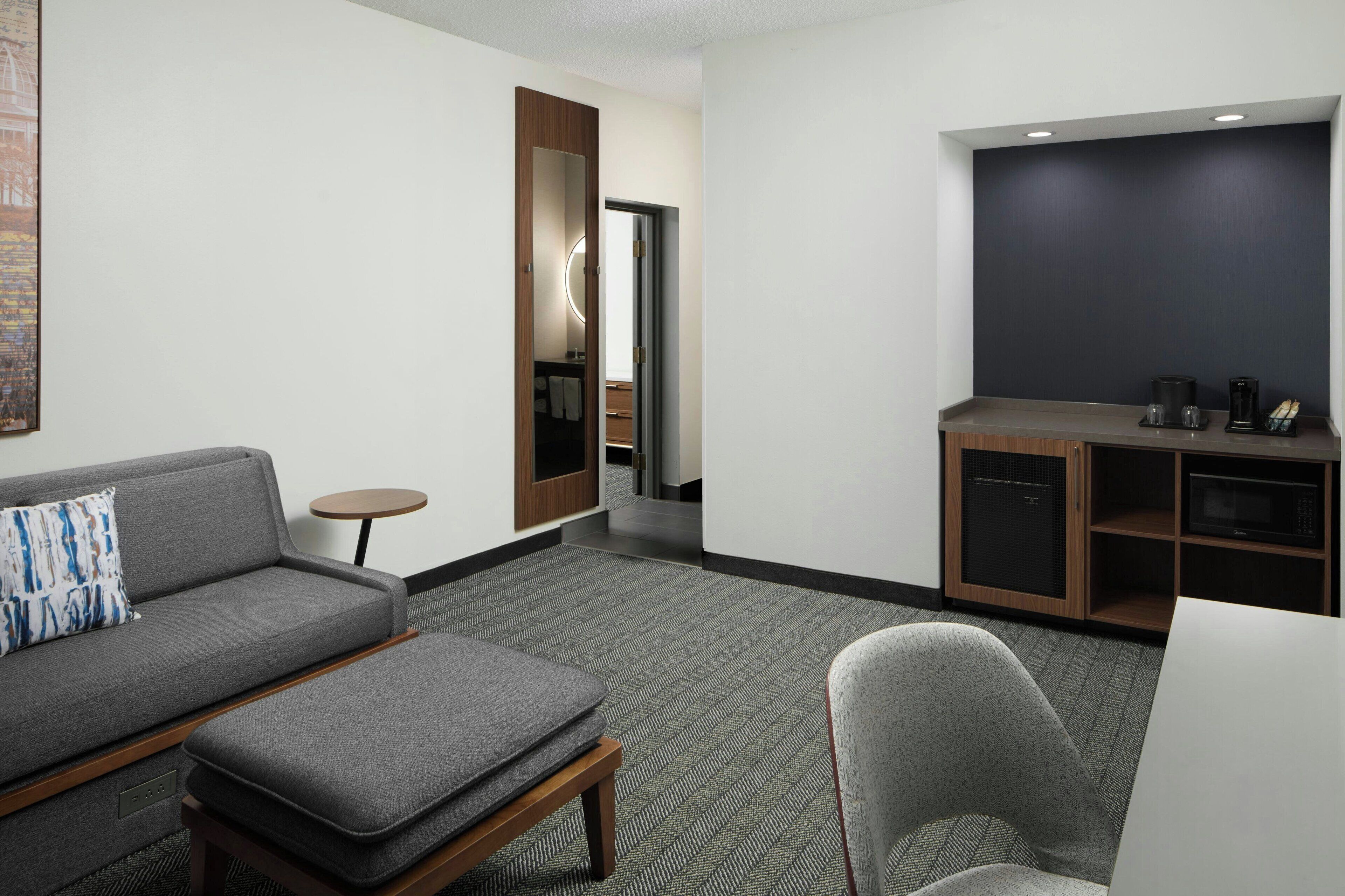 Ein modernes Hotelzimmer mit grauem Sofa, einem kleinen runden Tisch, einem grauen Sessel und einer Küchenzeile mit Mikrowelle und Kaffeemaschine.