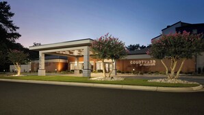 Un hotel Courtyard by Marriott con diseño moderno, que presenta una entrada cubierta y áreas ajardinadas con árboles y arbustos.