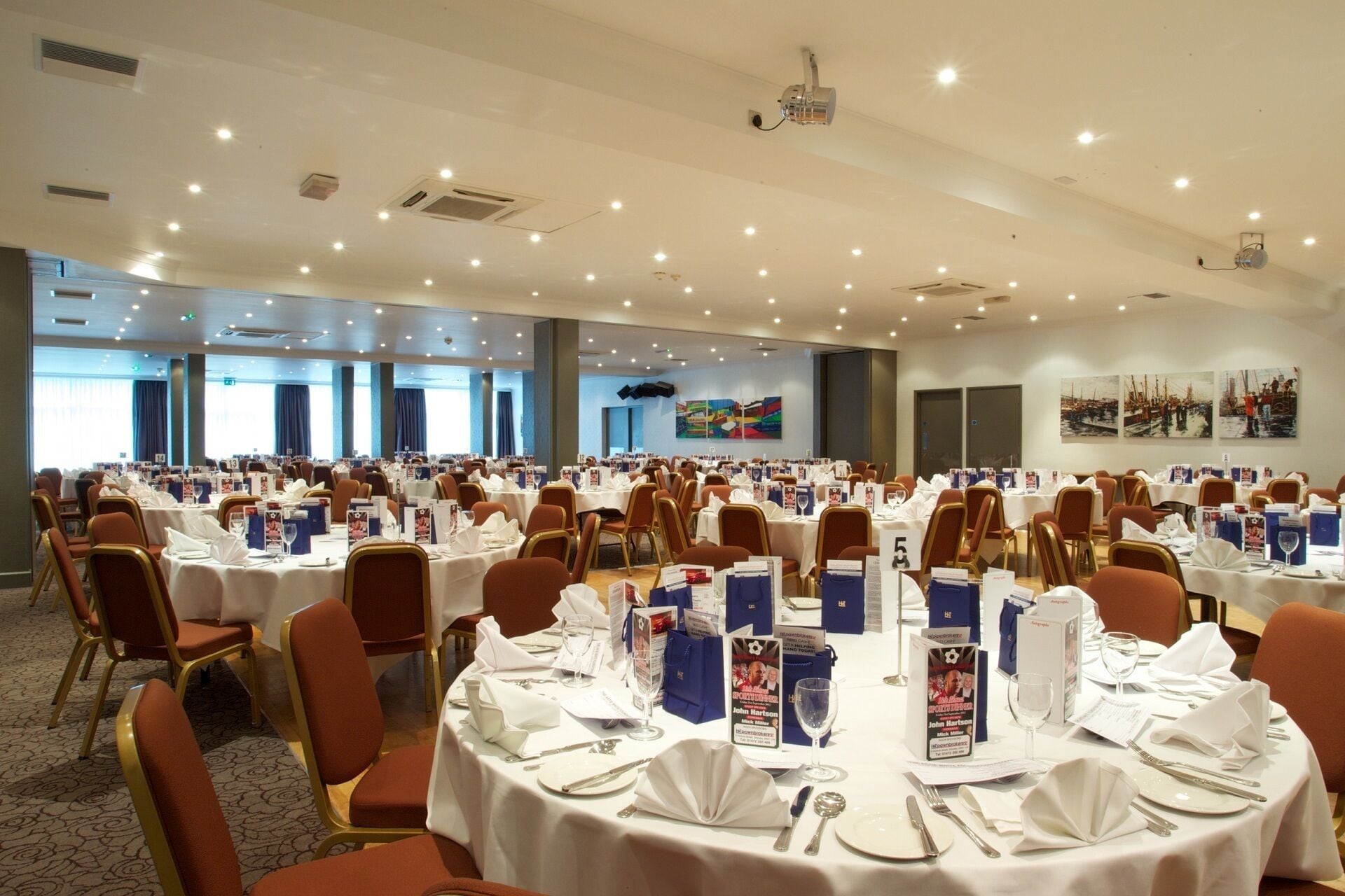 A large conference room with round tables, white tablecloths, and orange chairs.