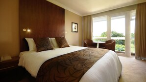 Habitación de hotel con una cama grande, una mesita y una silla. Se ve una ventana con cortinas y vistas a zonas verdes.