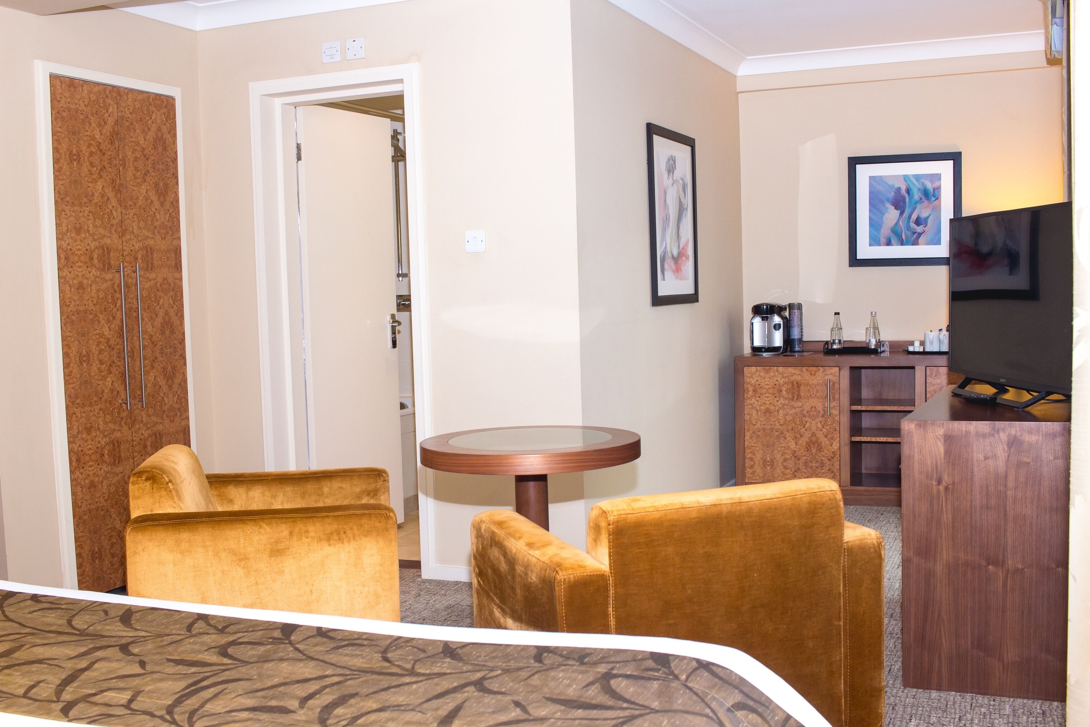 Una habitación de hotel con cama, dos sillones, una mesa redonda, minibar y obras de arte en las paredes.