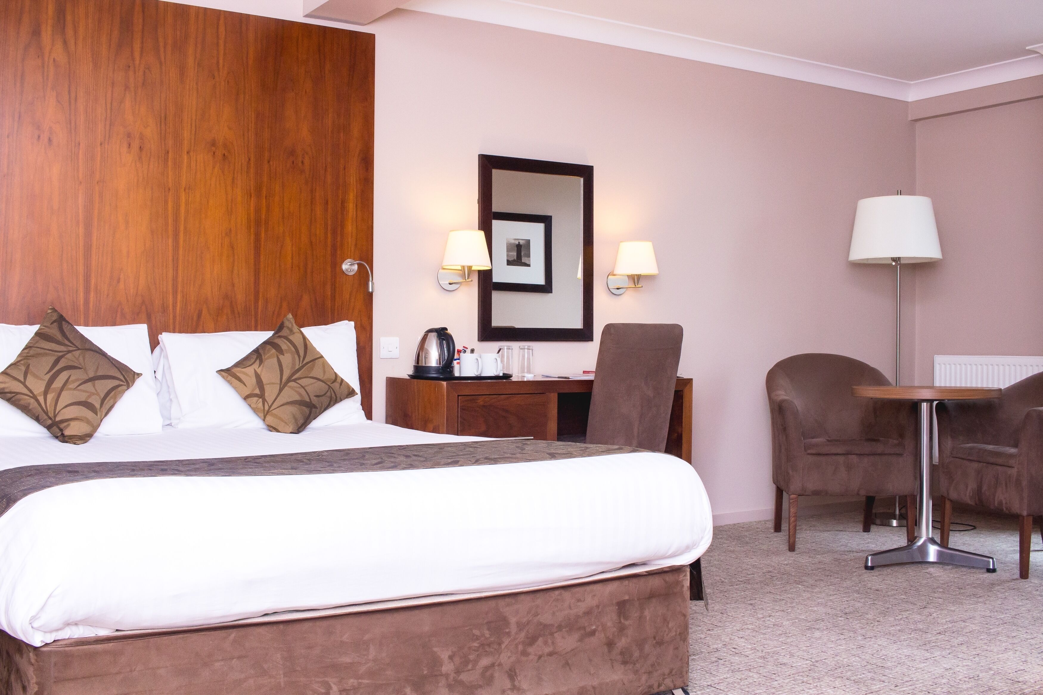 Habitación de hotel con una cama grande, cabecera de madera, un escritorio con silla, una lámpara y dos lámparas de pared.