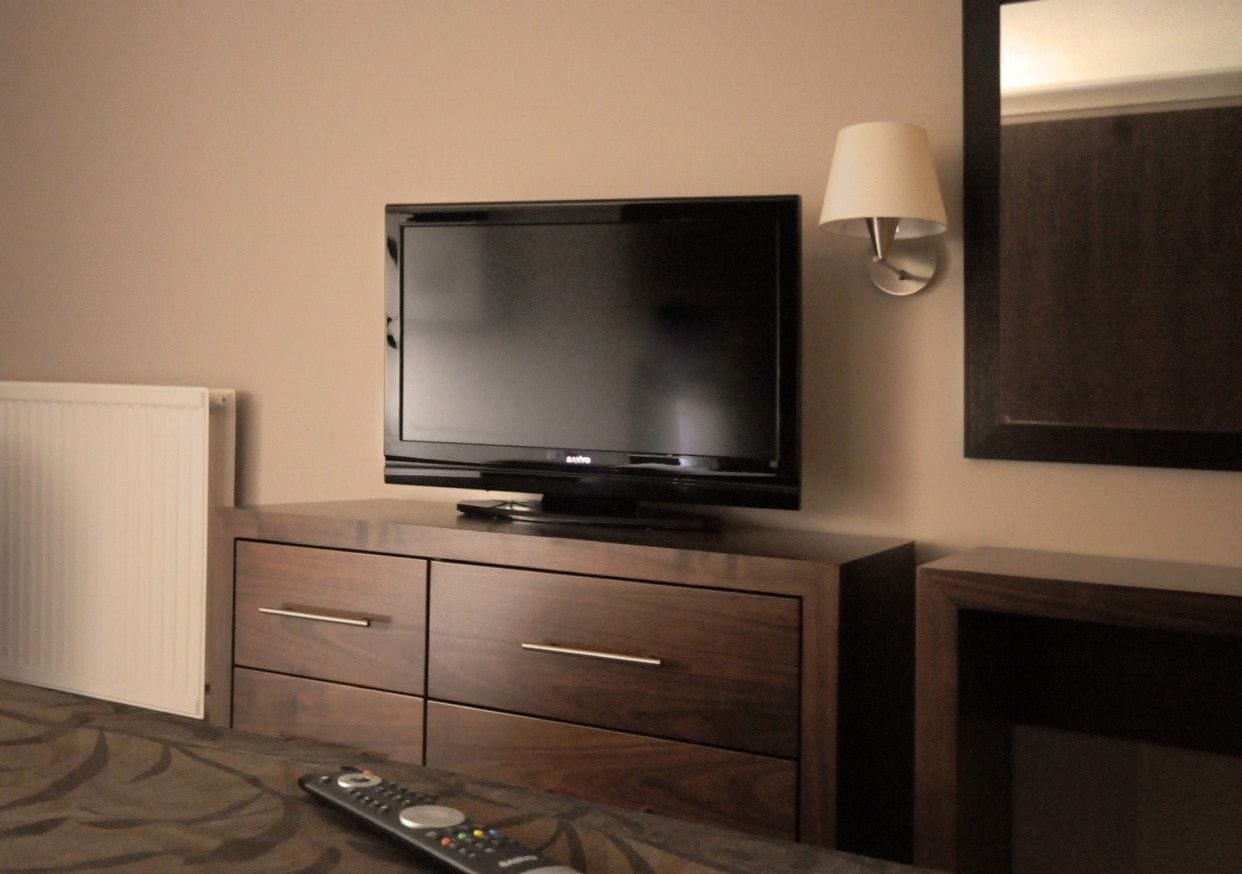 Un televisor sobre un mueble de madera con dos cajones, un control remoto y una lámpara fijada a la pared.