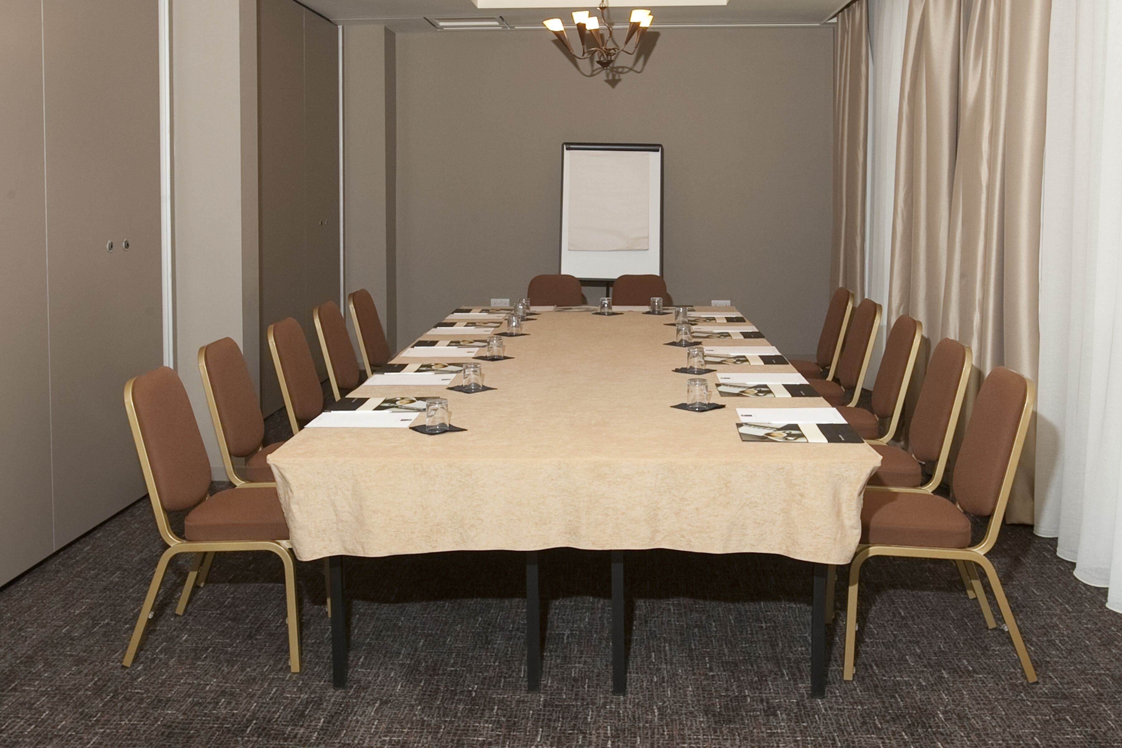 Een congreszaal met een lange tafel, bruine stoelen, een projectiescherm en een kroonluchter.