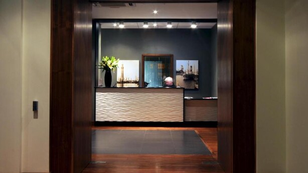 Un área de recepción moderna con suelo de madera, un mostrador de recepción, una planta en maceta y una pared azul con textura.