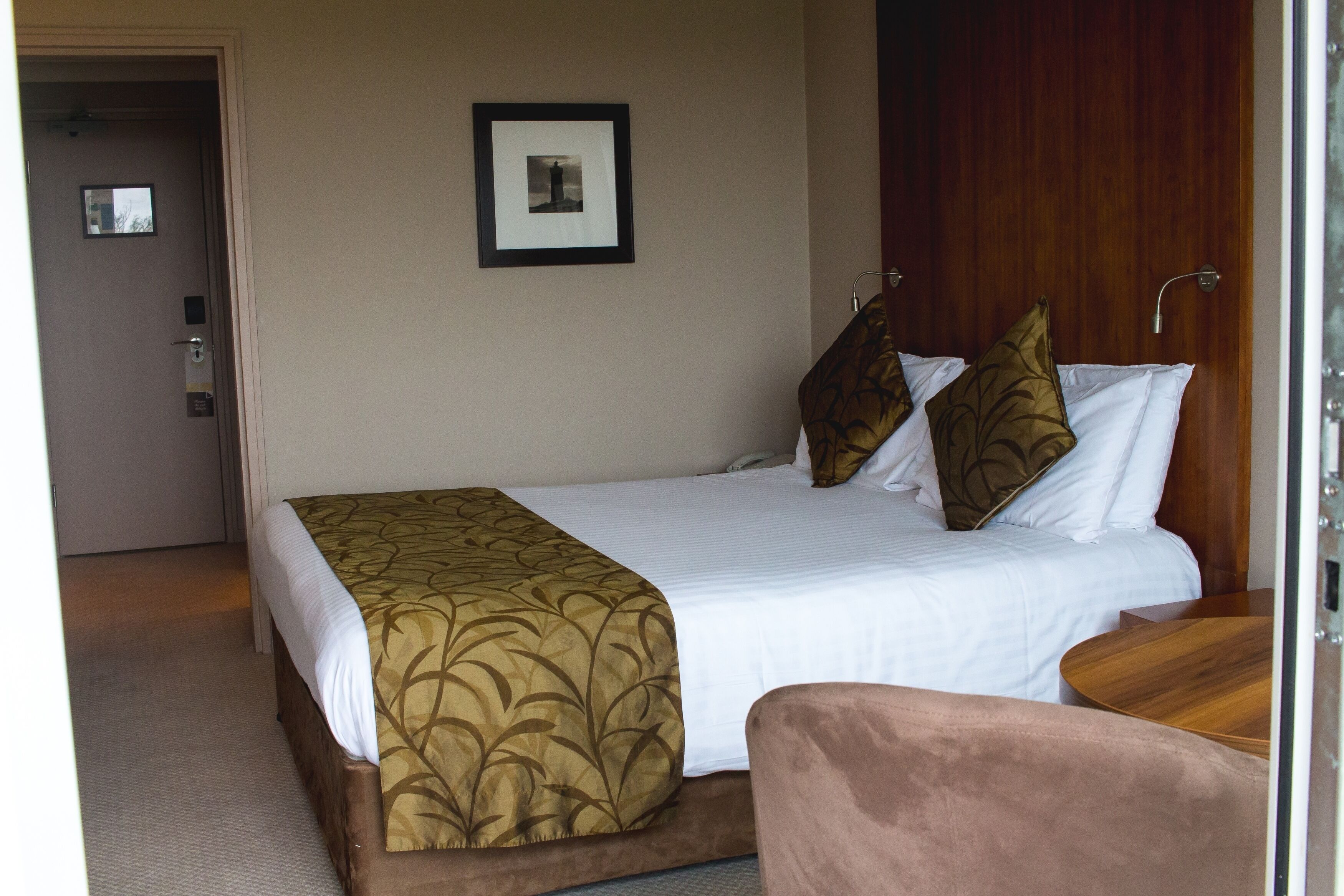 A hotel room with a bed, a wooden headboard, a framed picture on the wall, and a door leading to another room.