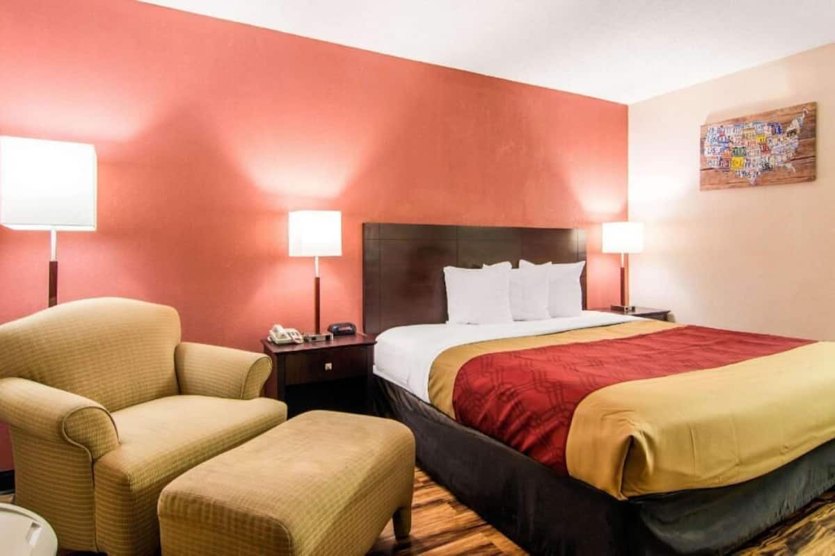A hotel room with a bed, two bedside lamps, a chair, a small table, and a wall art piece.