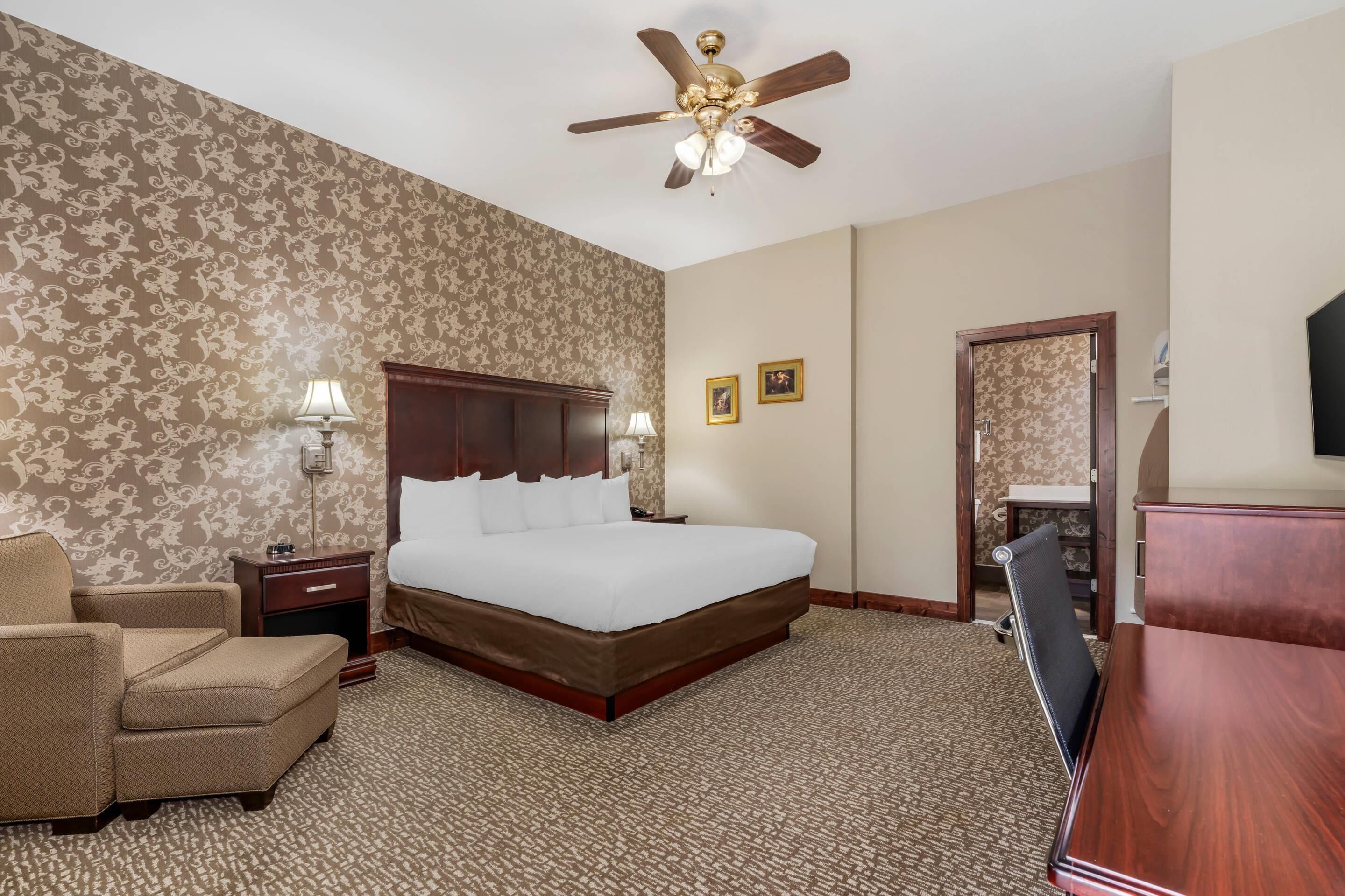 A hotel room with a large bed, a desk, a chair, a ceiling fan, and two wall-mounted lamps.
