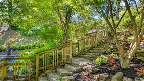 Eine steinerne Treppe mit hölzernen Geländern führt einen Hügel hinauf, umgeben von Grünpflanzen und einem kleinen Wasserspiel.