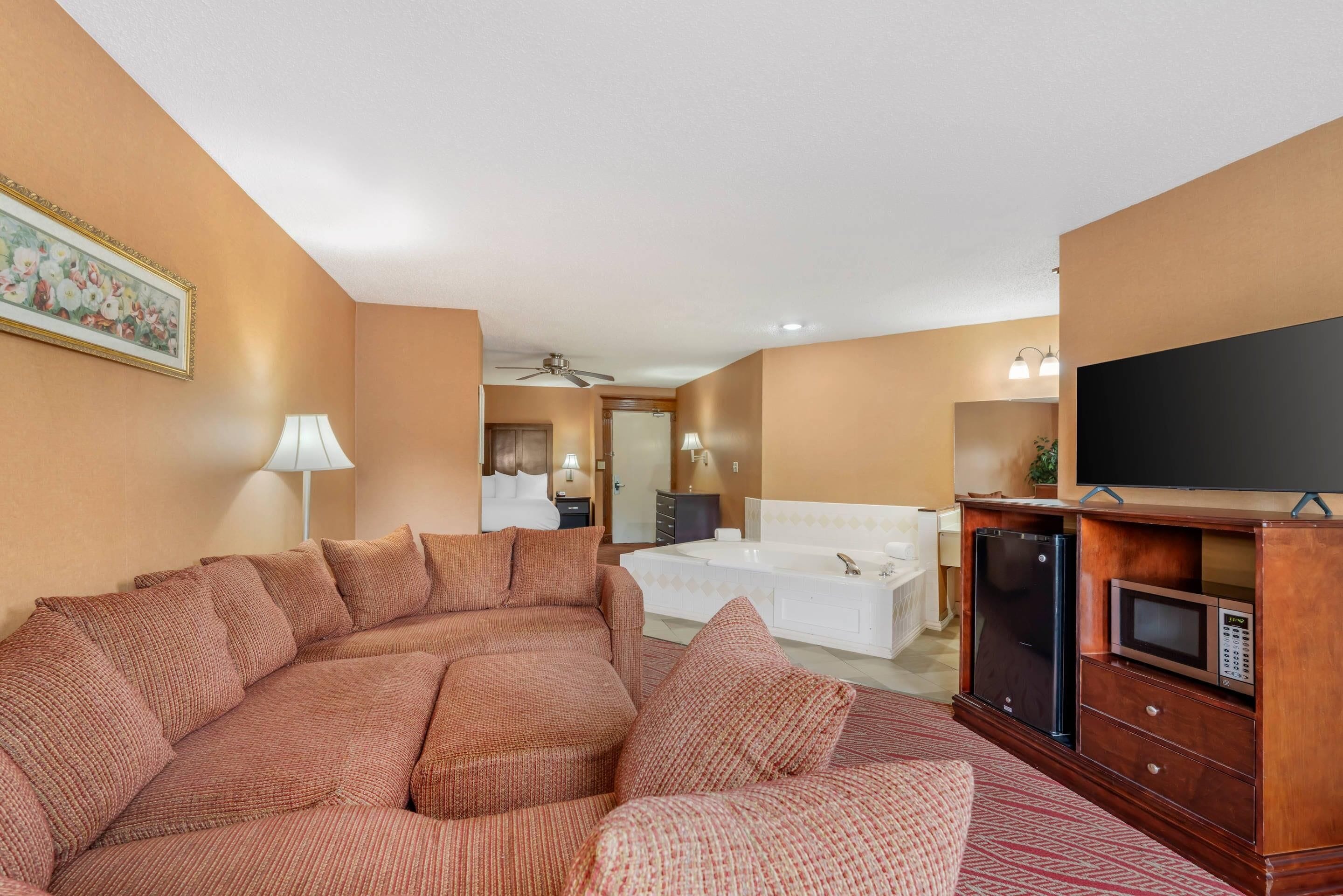 A hotel room with a large sectional sofa, a flat-screen TV, a microwave, a large window, and a bathroom with a bathtub and shower.