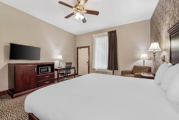 A hotel room with a large bed, a flat-screen TV, a desk with a chair, a microwave, a ceiling fan, and two lamps.