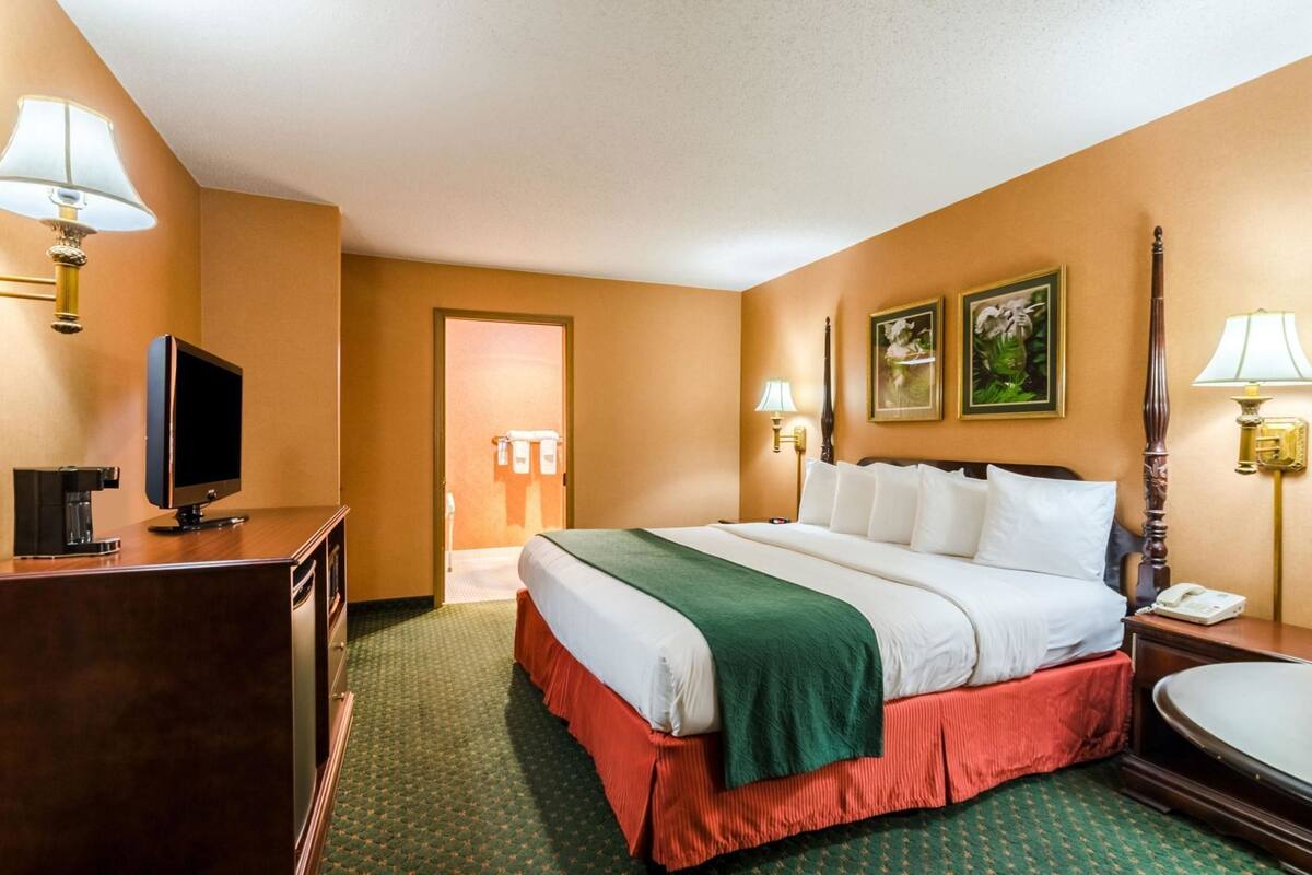 A hotel room with a bed, a desk with a television, a bedside table with a lamp, and a bathroom door.