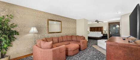 A hotel room with a sectional sofa, a desk, a television, a ceiling fan, a bed, and a potted plant.