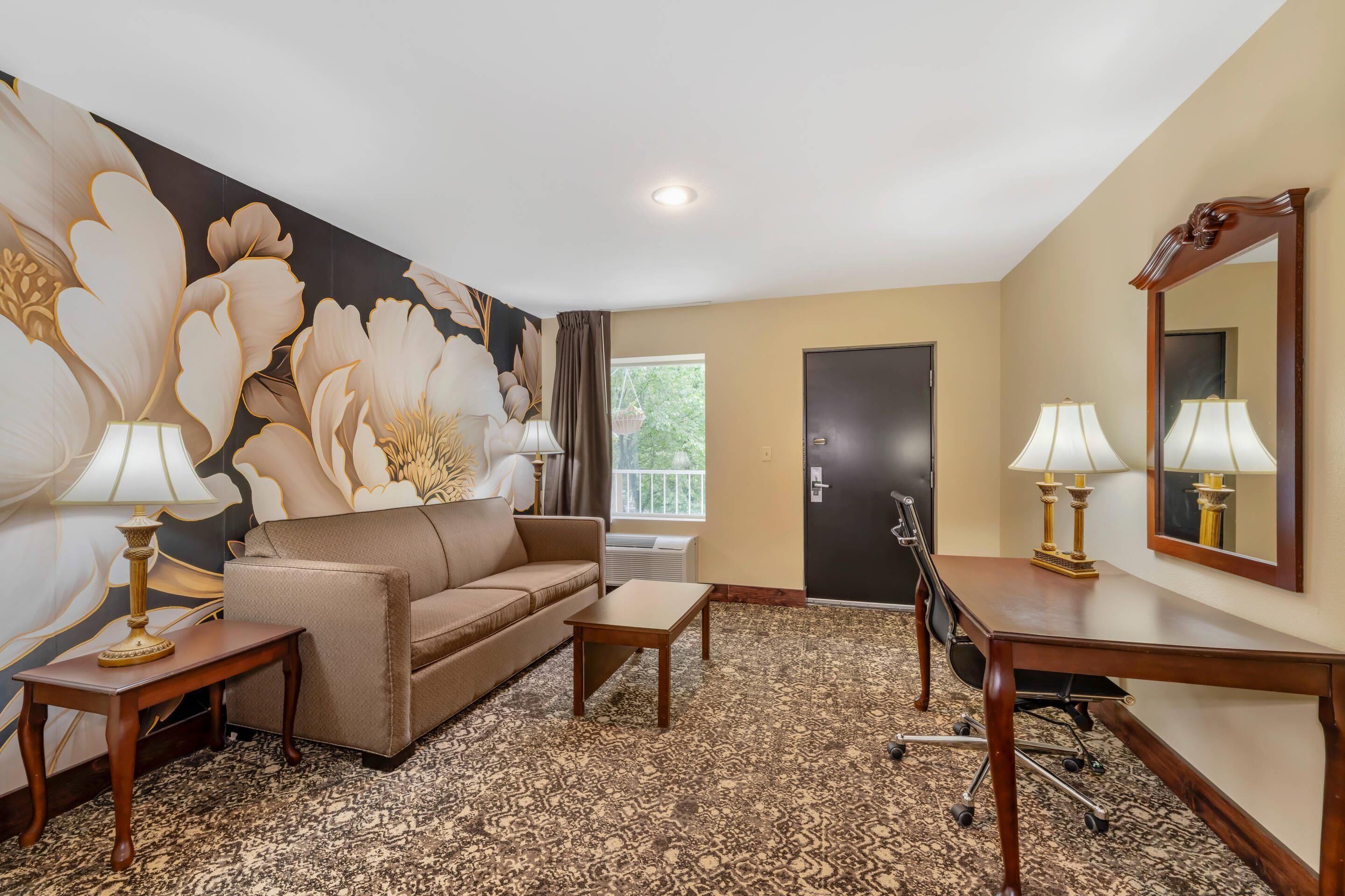 A hotel room with a sofa, two tables, a desk, a large mirror, and a floral wall mural.