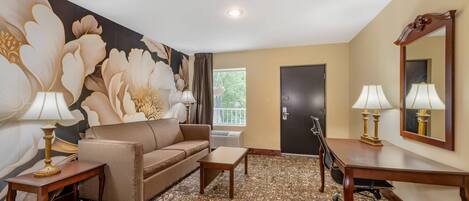 A hotel room with a sofa, two tables, a desk, a large mirror, and a floral wall mural.
