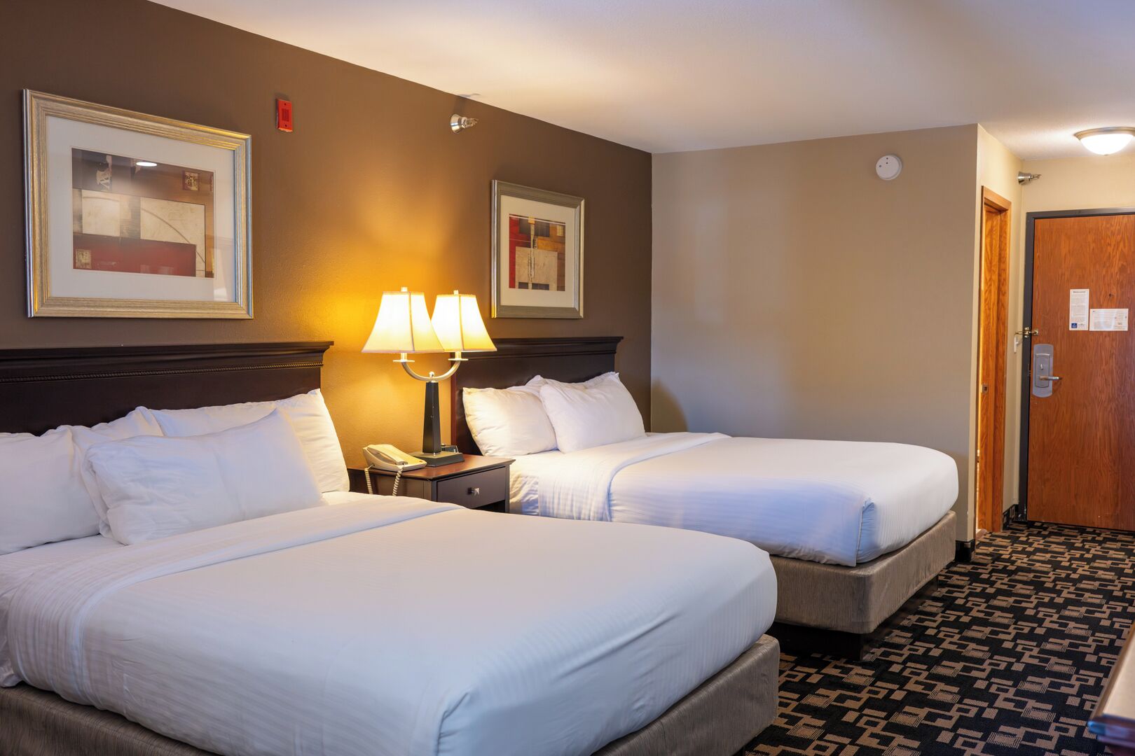 A hotel room with two beds, a nightstand with a lamp, a framed picture on the wall, and a door with a sign.