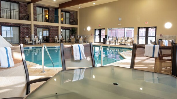 Un espace piscine d’hôtel comprenant plusieurs chaises longues, une piscine et un balcon au deuxième étage avec des fenêtres.