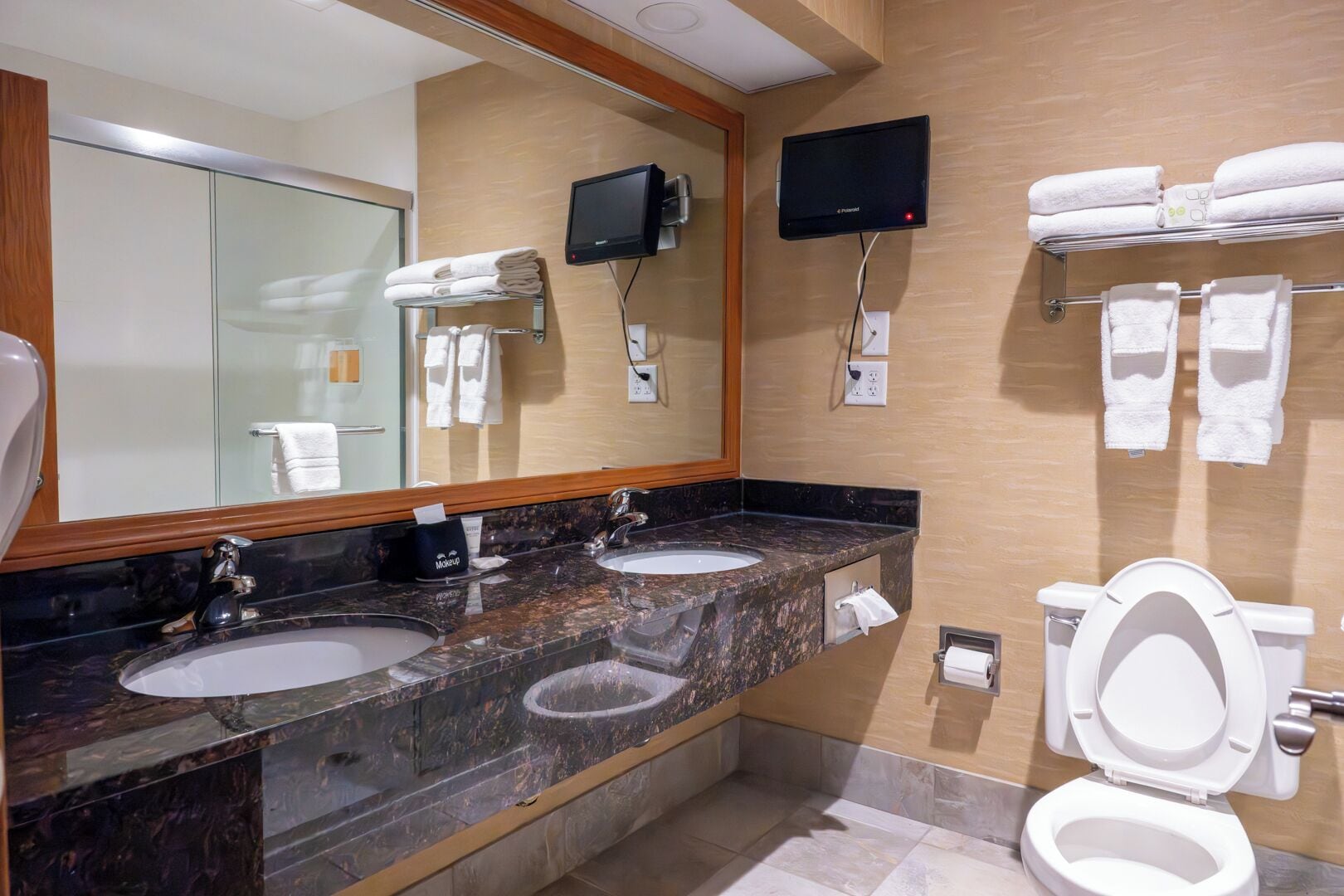 A hotel bathroom with two sinks, a toilet, a TV mounted on the wall, and a towel rack with towels.