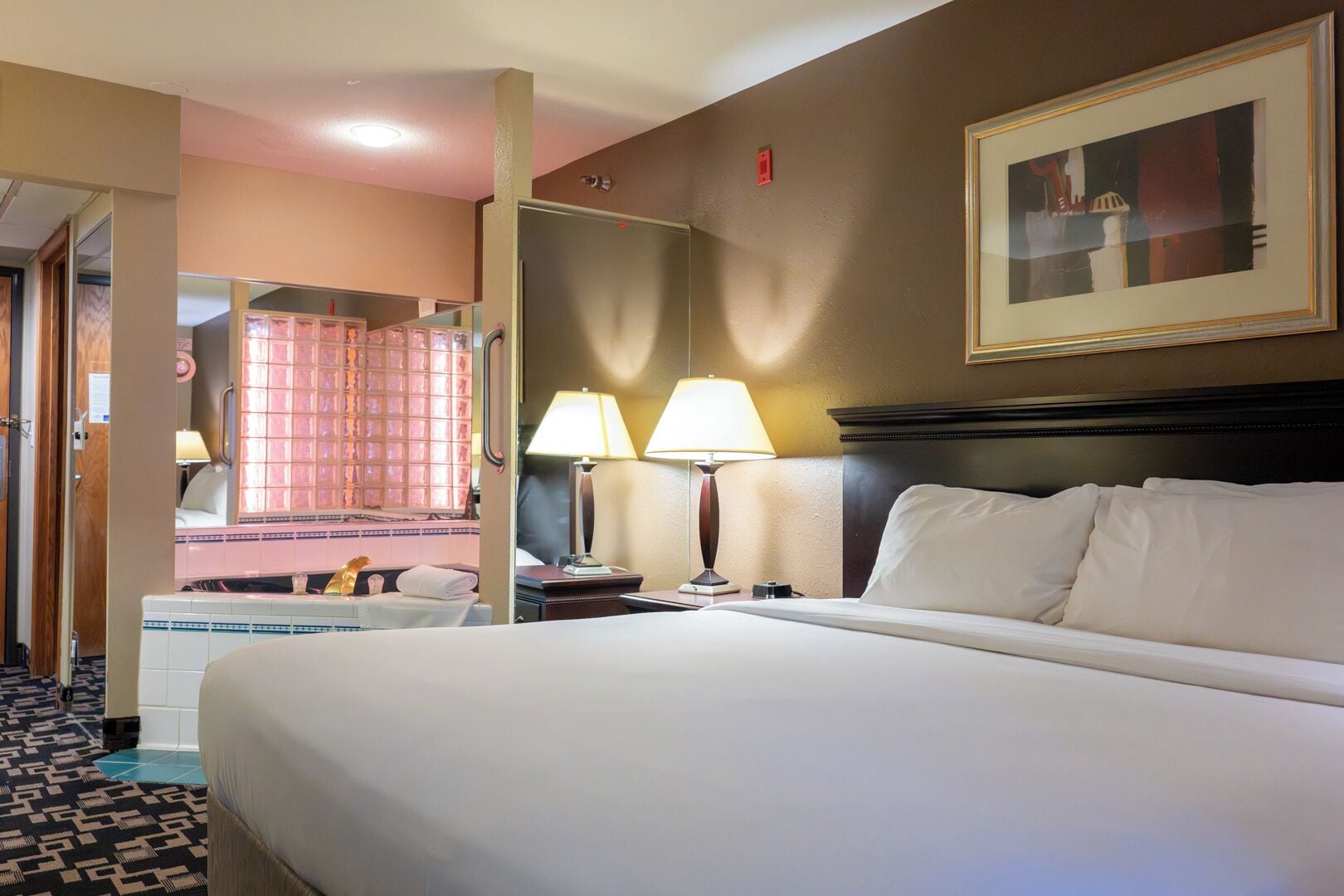 Une chambre d’hôtel avec un lit, deux lampes de chevet, une table de nuit, un miroir, une fenêtre avec des rideaux roses et un tableau encadré au mur.