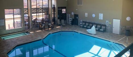 An indoor swimming pool with clear pool rules and spa rules signs, a covered seating area, and a view of the outside area through large windows.