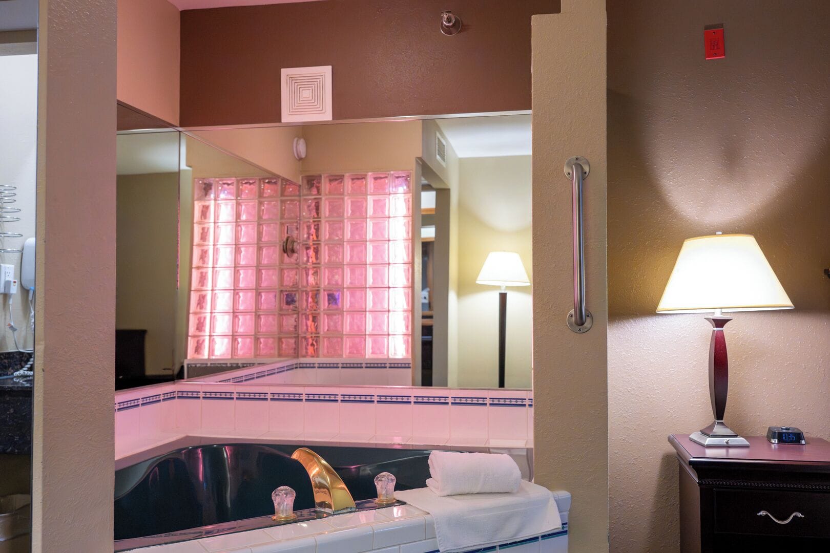 A hotel room with a large mirror, a bathtub, a bedside table with a lamp, and a clock.