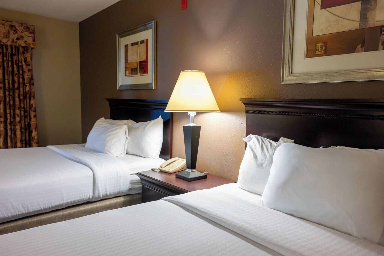 A hotel room with two beds, a nightstand with a lamp, and framed artwork on the walls.