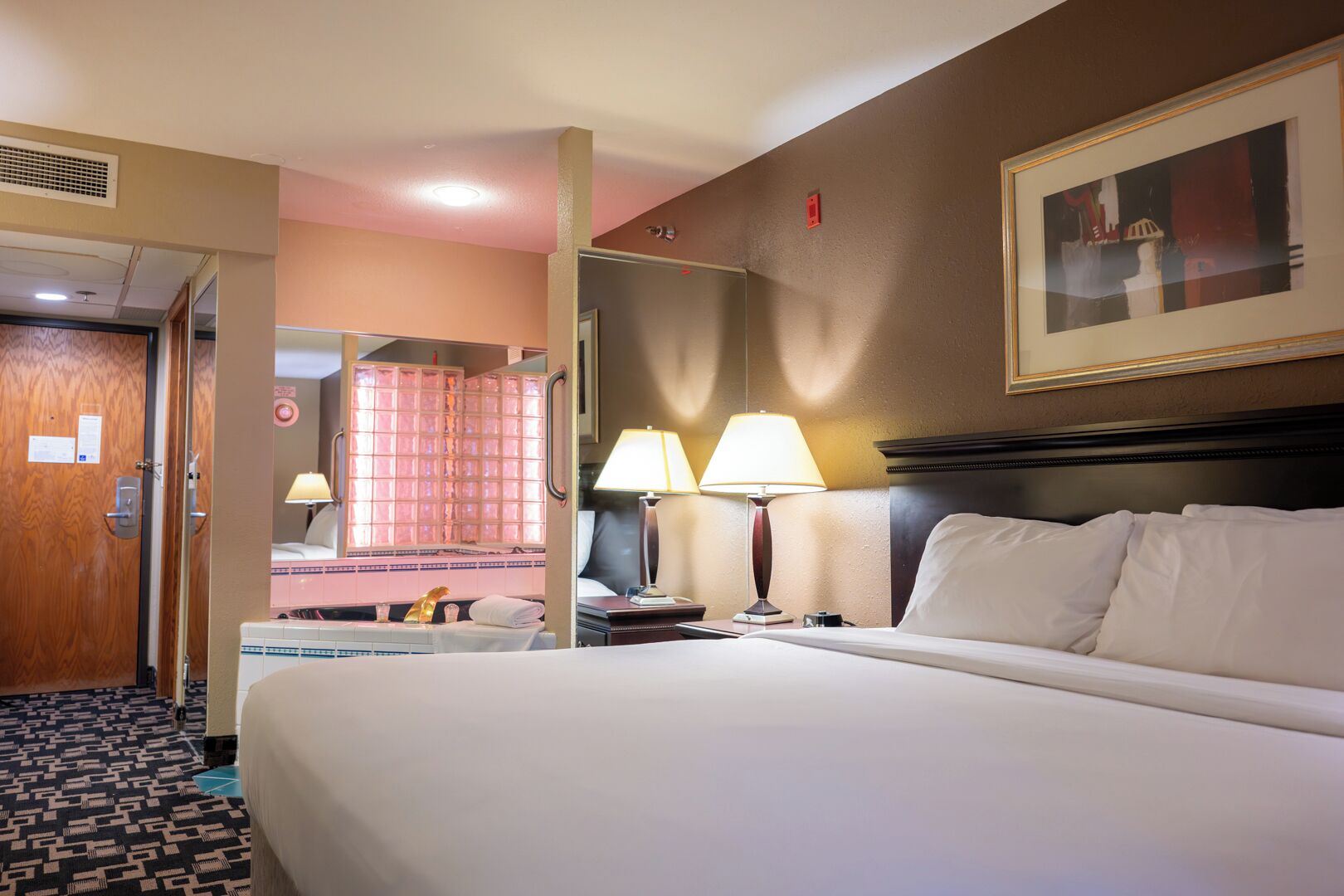 A hotel room with a large bed, two bedside lamps, a nightstand, a mirror, a door, and a framed picture on the wall.