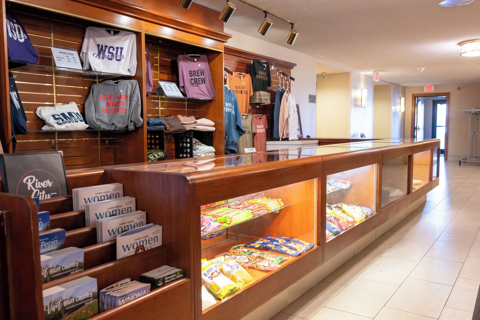 Zona comercial con estanterías de ropa y libros, una vitrina con aperitivos y una barra de madera.