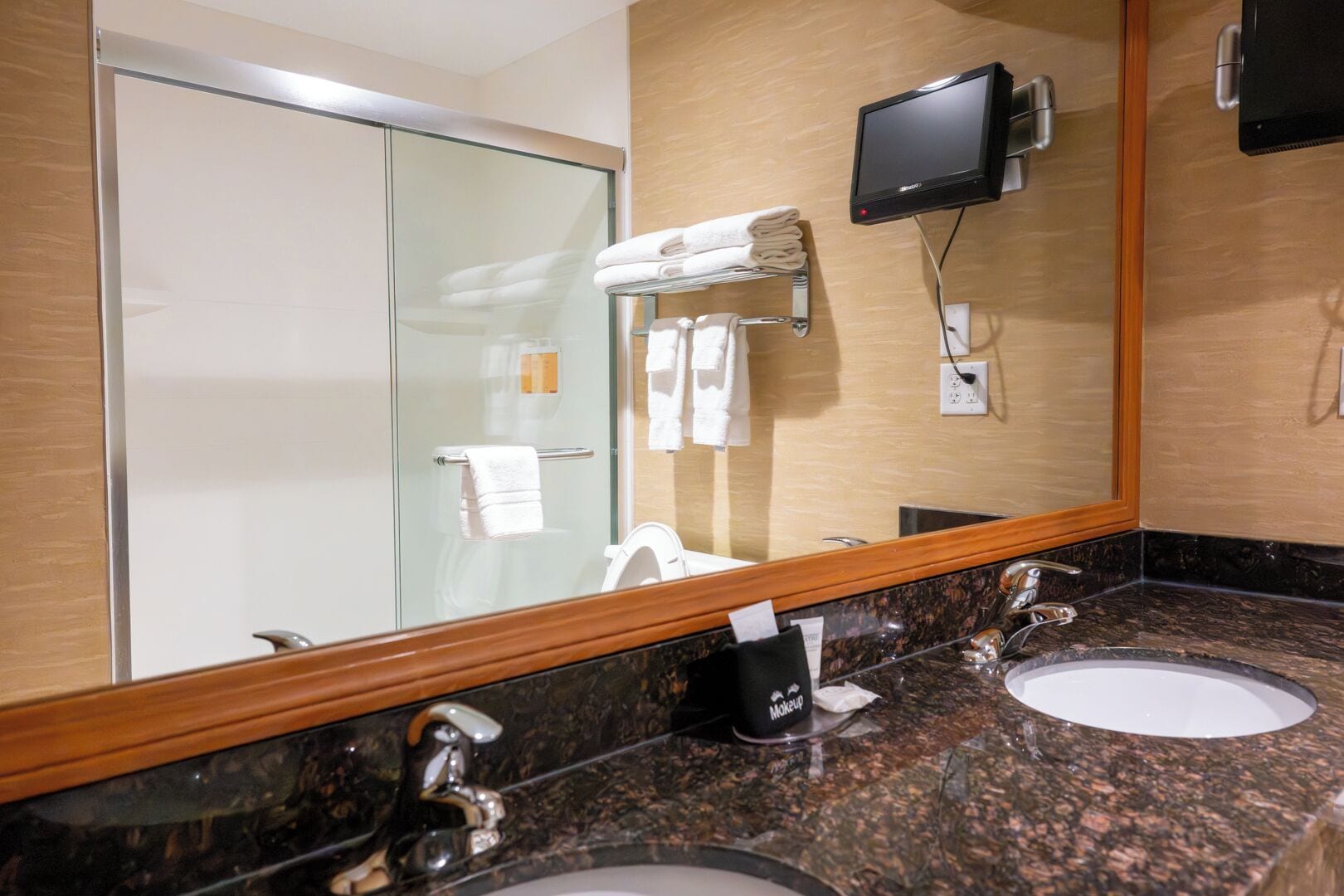 Un bagno d'hotel con cabina doccia in vetro, due lavabi con specchiette a muro, una televisione montata e un portaservizi con asciugamani piegati.