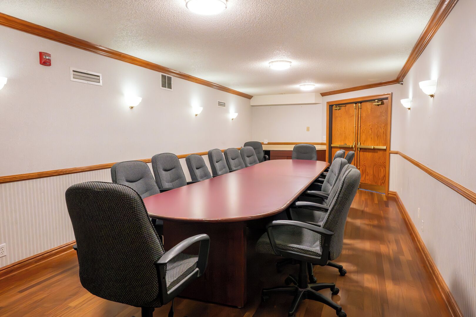 Un salón de conferencias con una larga mesa, varias sillas, una puerta de madera y luces fijadas a la pared.