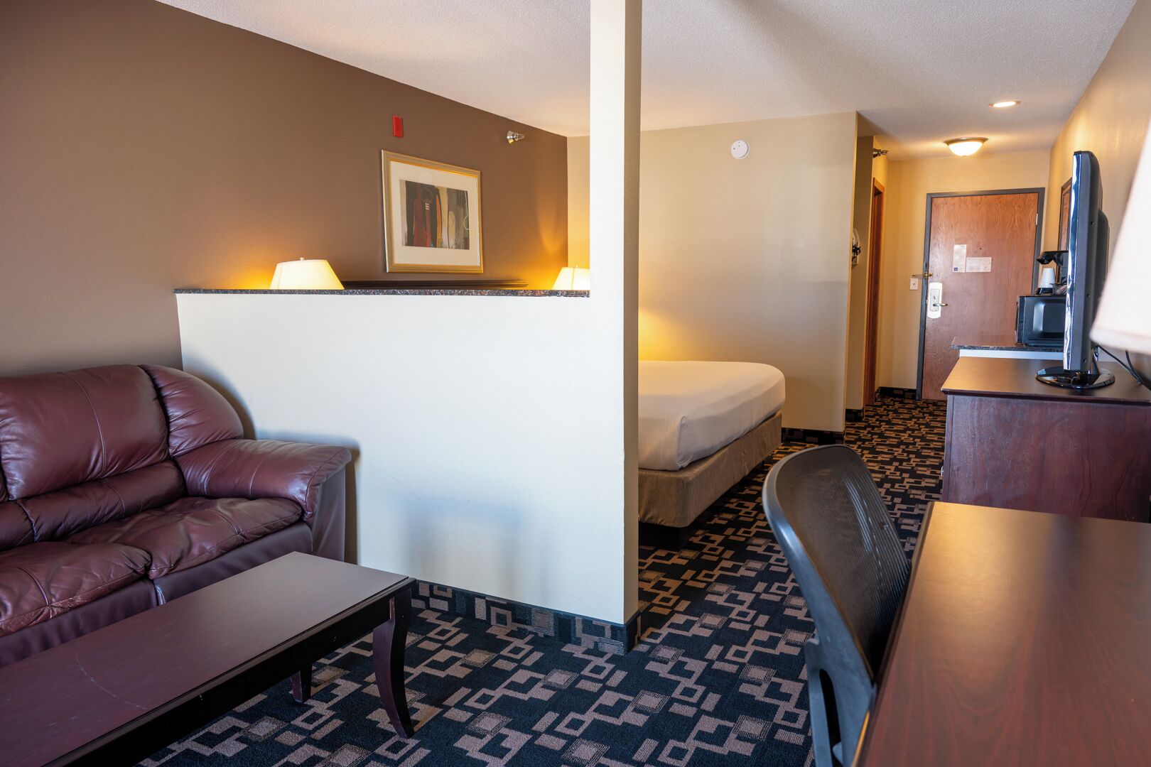A hotel room with a leather sofa, a small table, a bed, a desk with a computer, and a television.