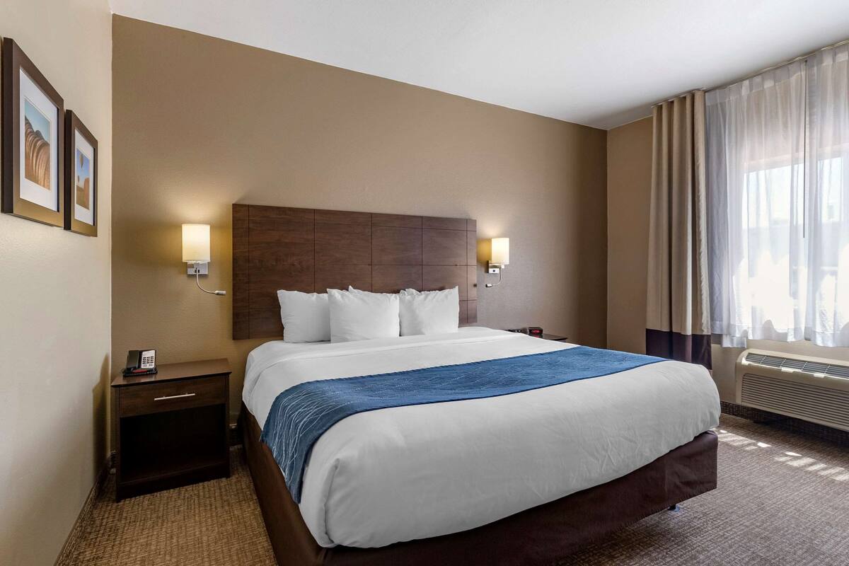 Une chambre d’hôtel avec un grand lit, deux tables de chevet, une petite table, un téléphone fixé au mur et une fenêtre avec des rideaux.
