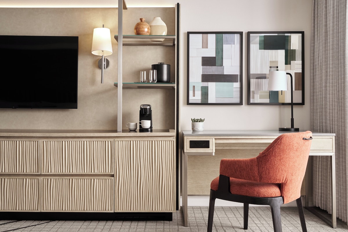 A modern hotel room with a flat-screen TV, a desk with a chair, a coffee machine, and a shelf with decorative items.