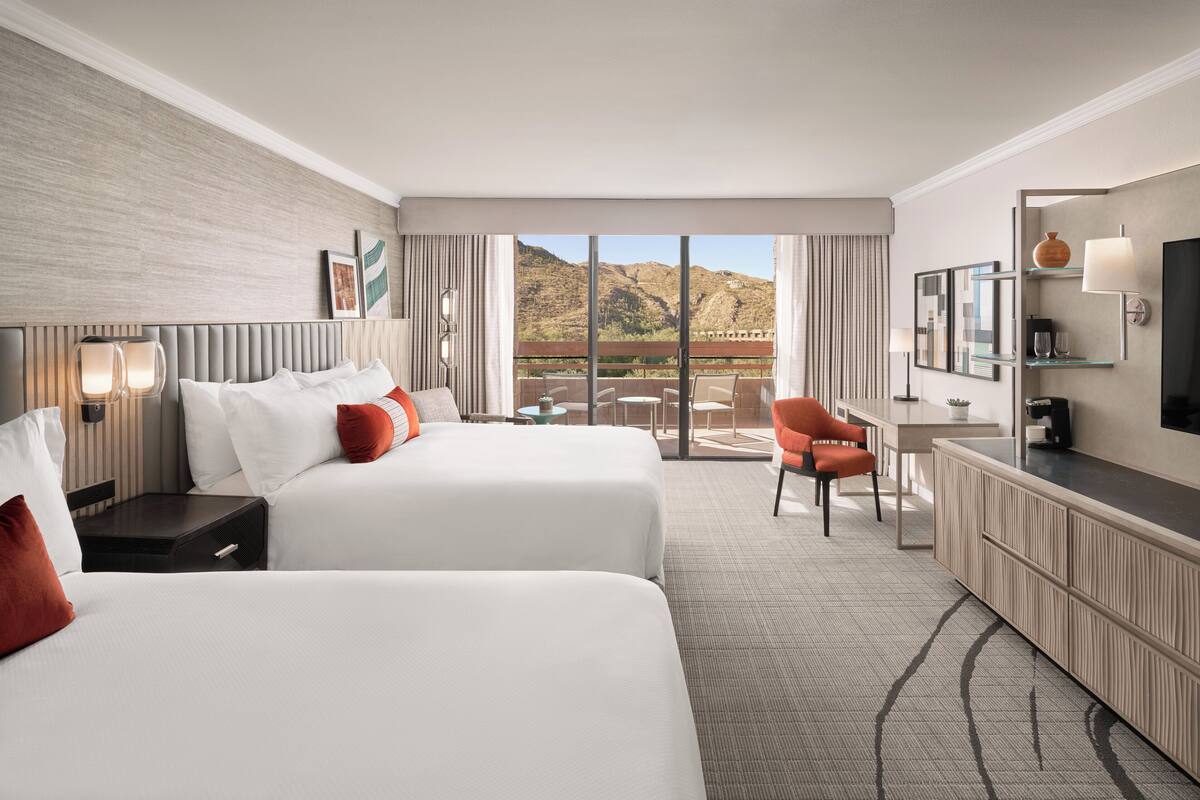 A hotel room with two beds, a desk, a chair, a TV, and a balcony with a mountain view.