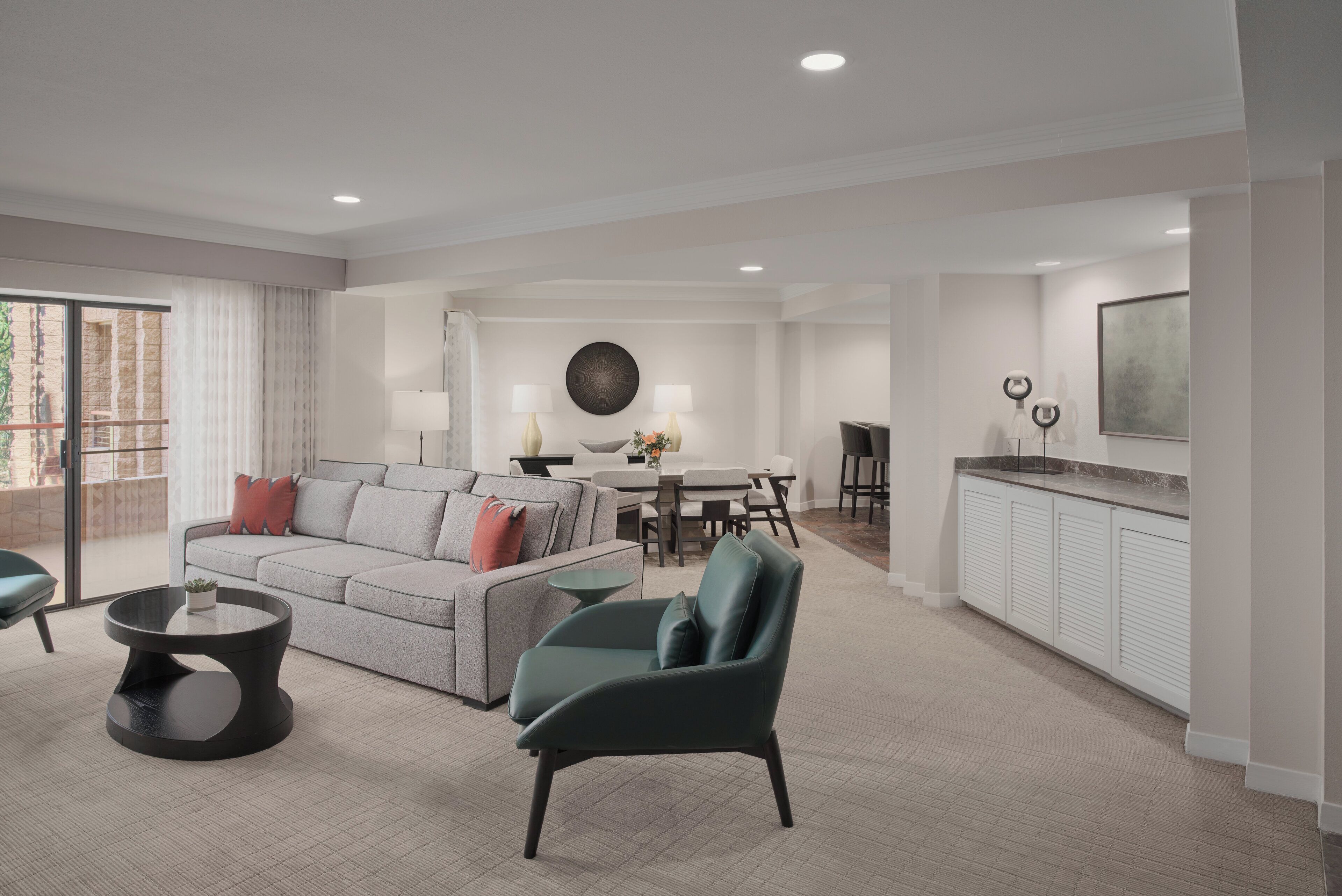 Un salon moderne avec un canapé gris, un fauteuil turquoise et une table basse ronde. On y trouve également un coin repas avec une table et des chaises, ainsi qu’une cuisine équipée de placards et d’un plan de travail.