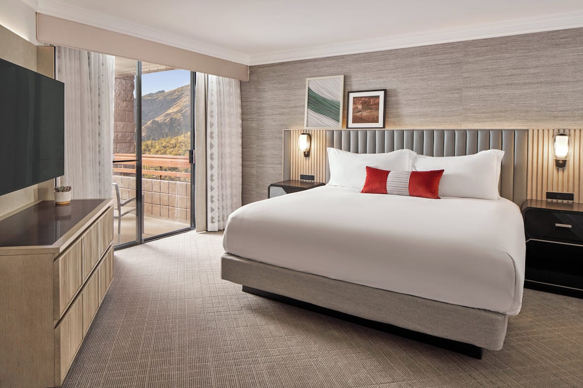 A hotel room with a large bed, a TV, a desk, and a view of the mountains through the sliding glass door.