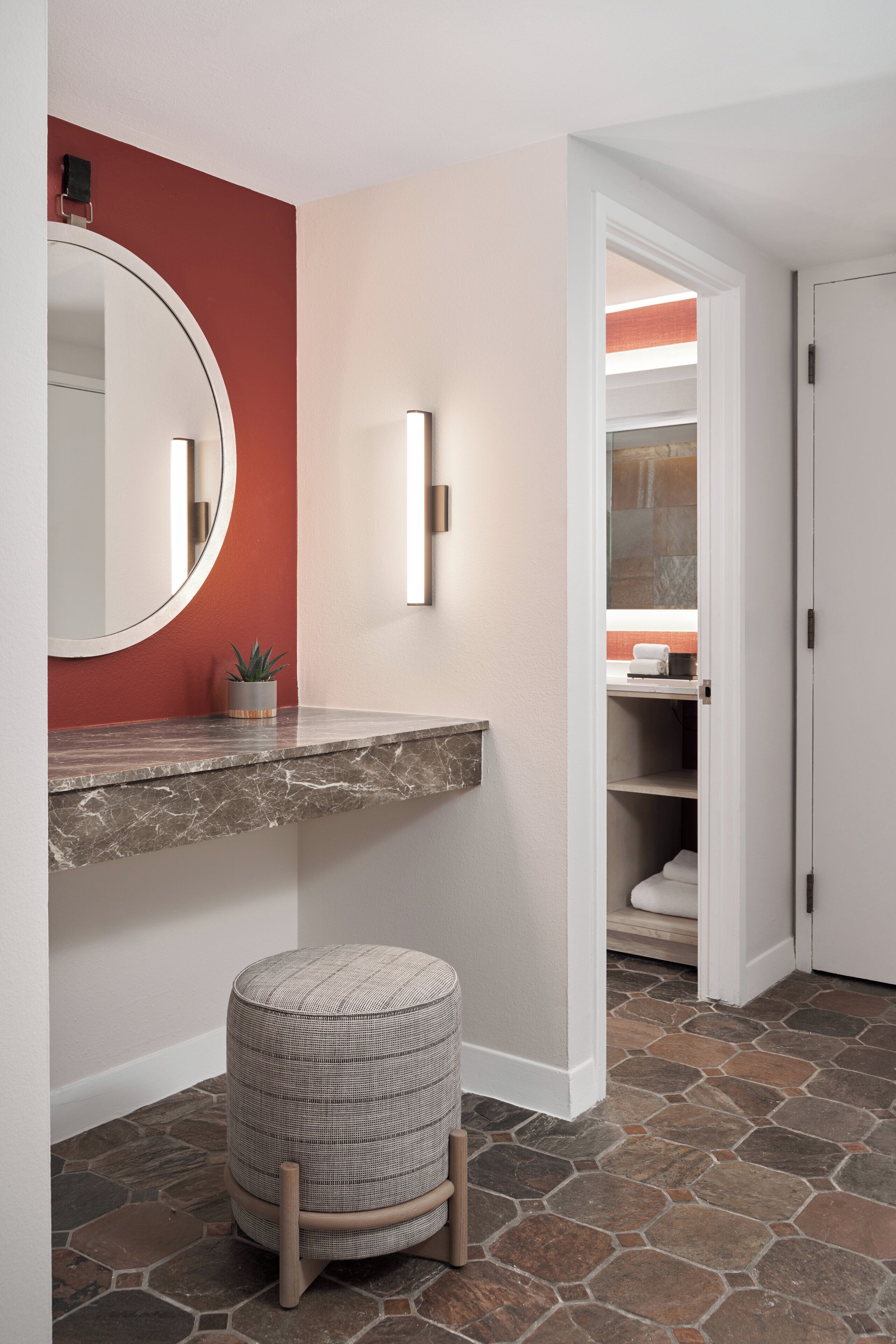 A bathroom with a marble countertop, a round mirror, a potted plant, and a stool.