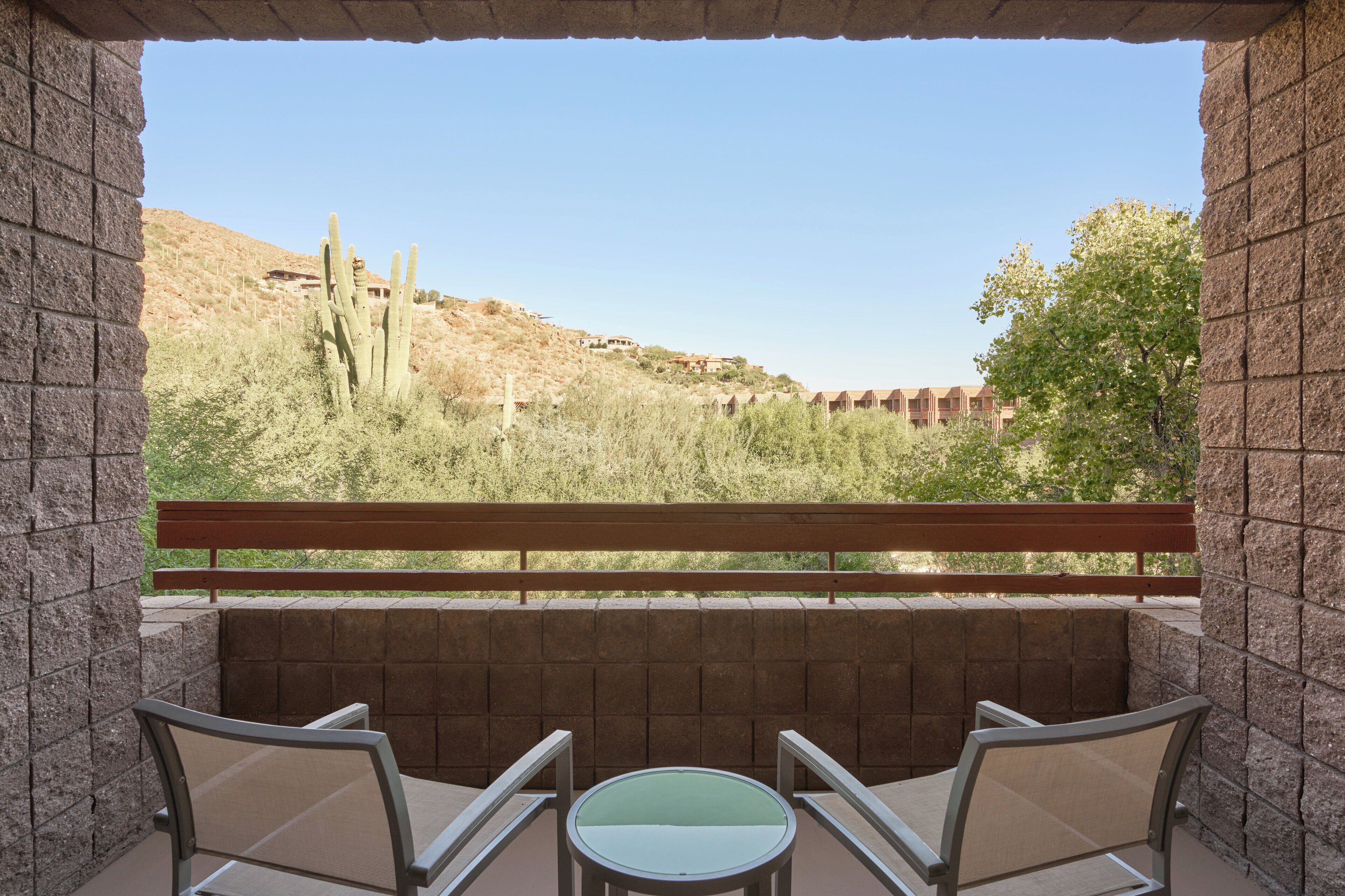 A balcony with two chairs and a table, overlooking a desert landscape with cacti and a distant building.