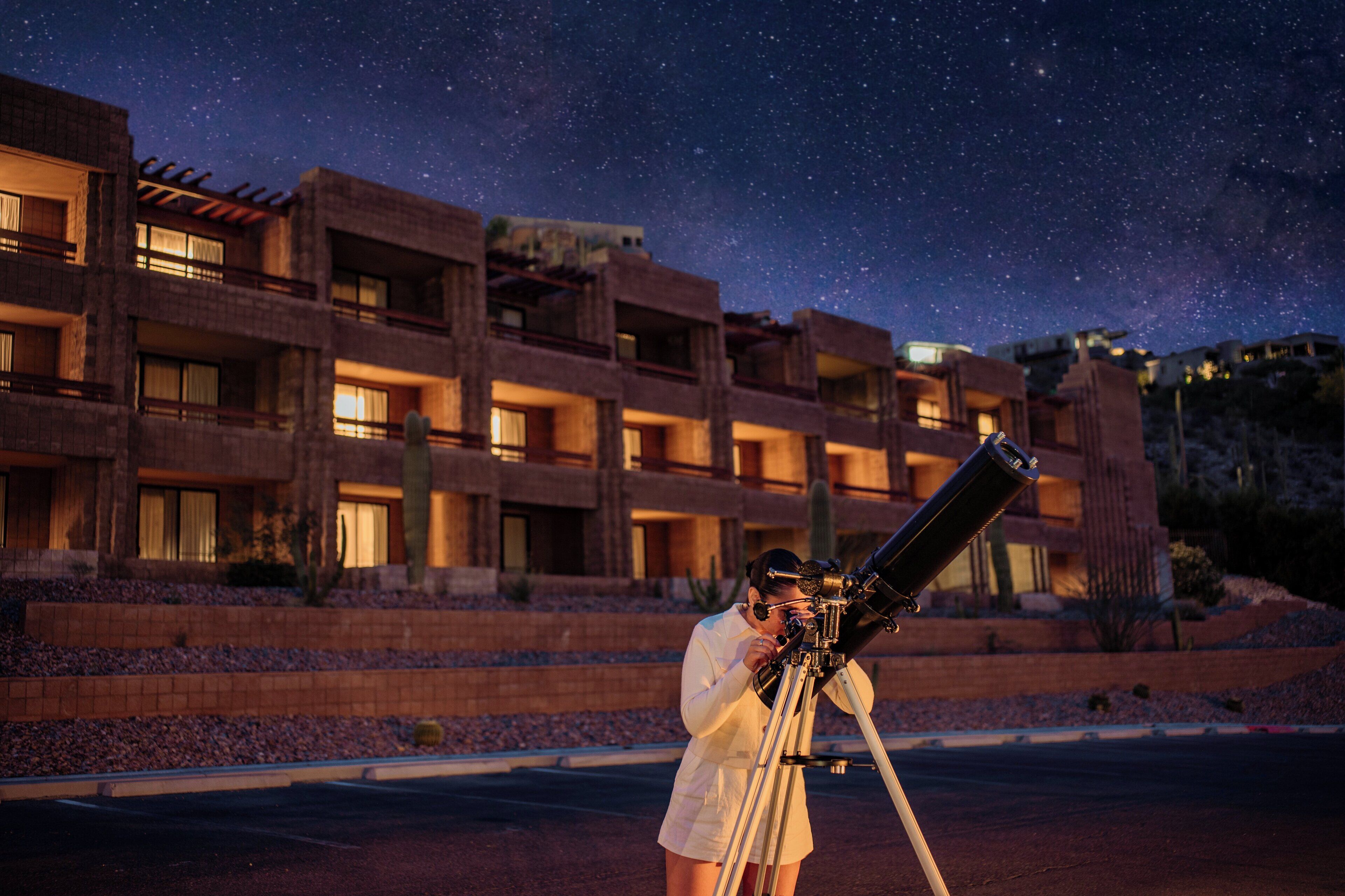 Una persona observant el cel estrellat amb un telescopi davant d'un edifici amb balcons múltiples.