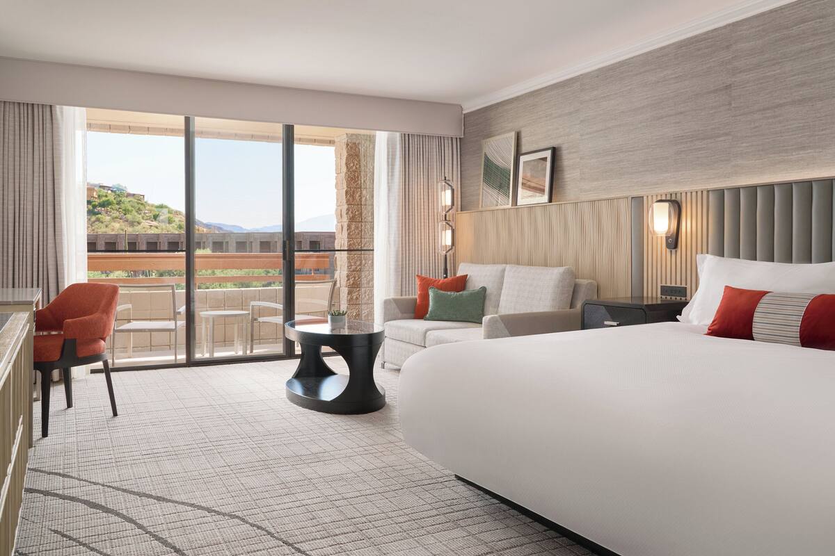 A hotel room with a large bed, a sofa, a small table, and a chair. There is a balcony with a view of a building and a hill.