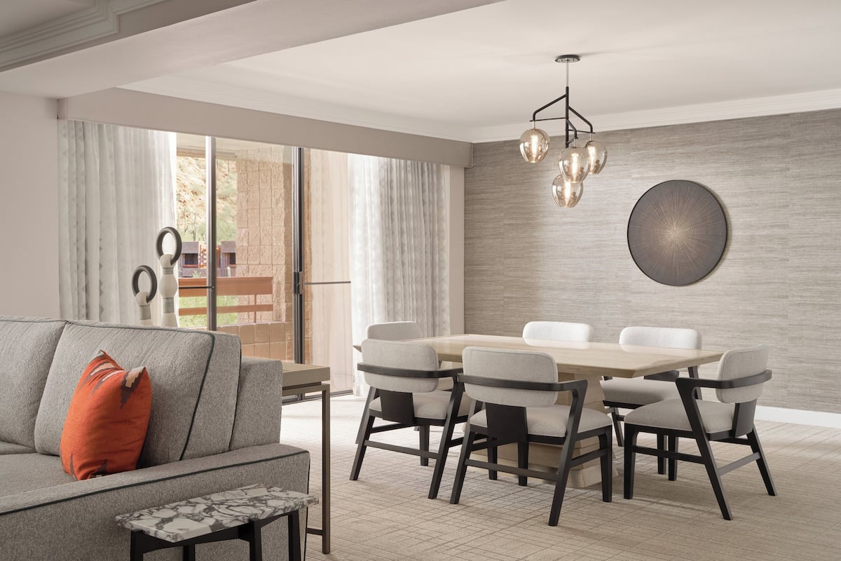 A modern dining area with a wooden table and chairs, a grey sofa with a red pillow, and a large window with curtains.