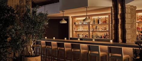 A bar area with wooden chairs, a large potted plant, and a well-lit bar counter with bottles.