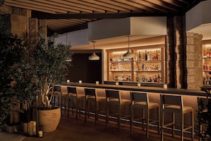Un área de bar con sillas de madera, una planta grande en maceta y una barra bien iluminada con botellas.