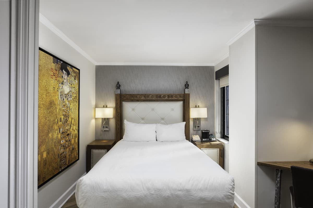 A hotel room with a large bed, two bedside tables with lamps, a large painting on the wall, and a desk with a chair.