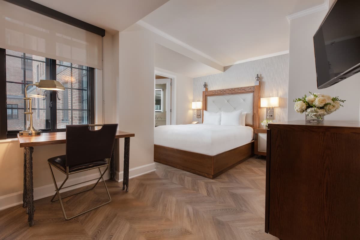 A hotel room with a large bed, a desk with a chair, a TV, and a window with blinds.
