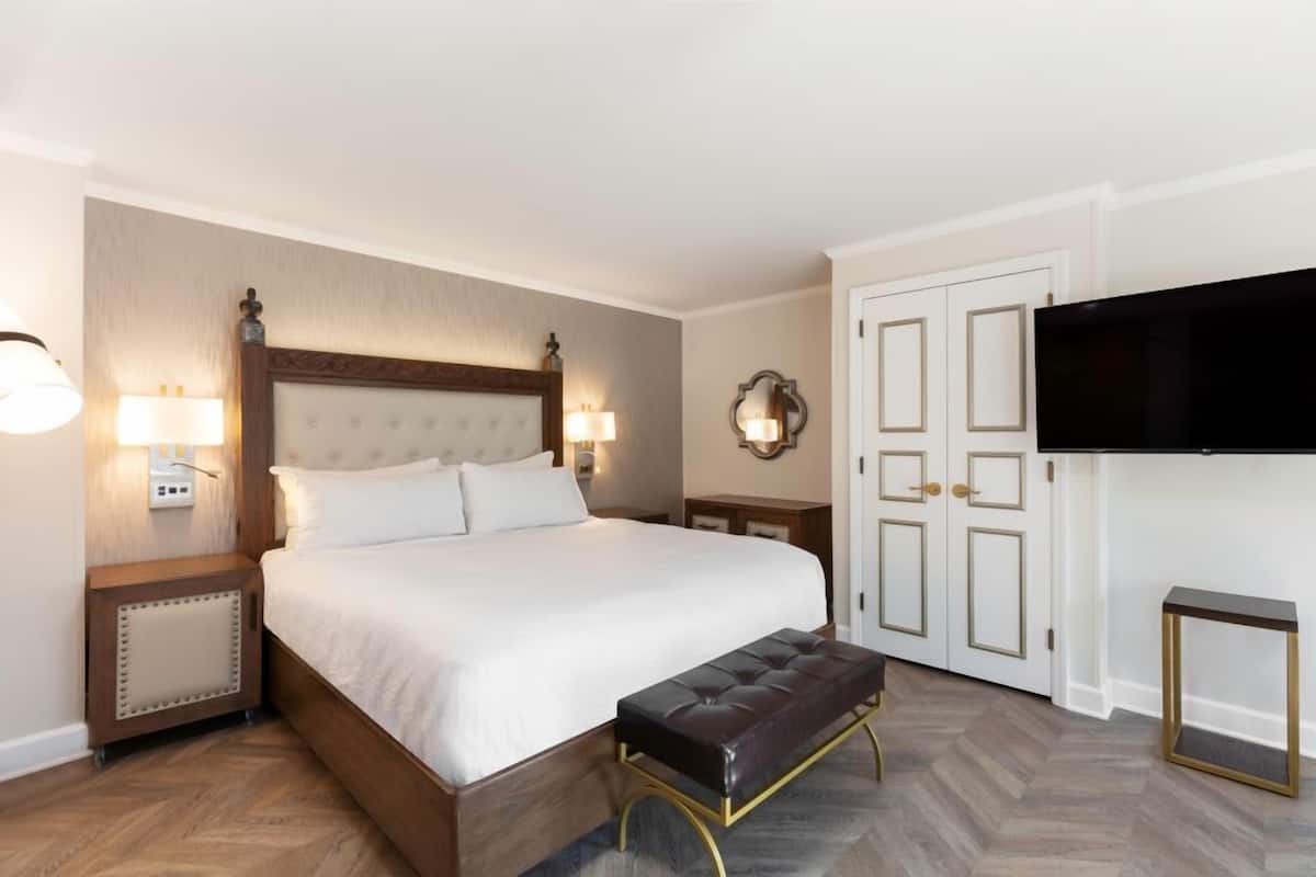 A hotel room with a large bed, bedside lamps, a television, and a bench.